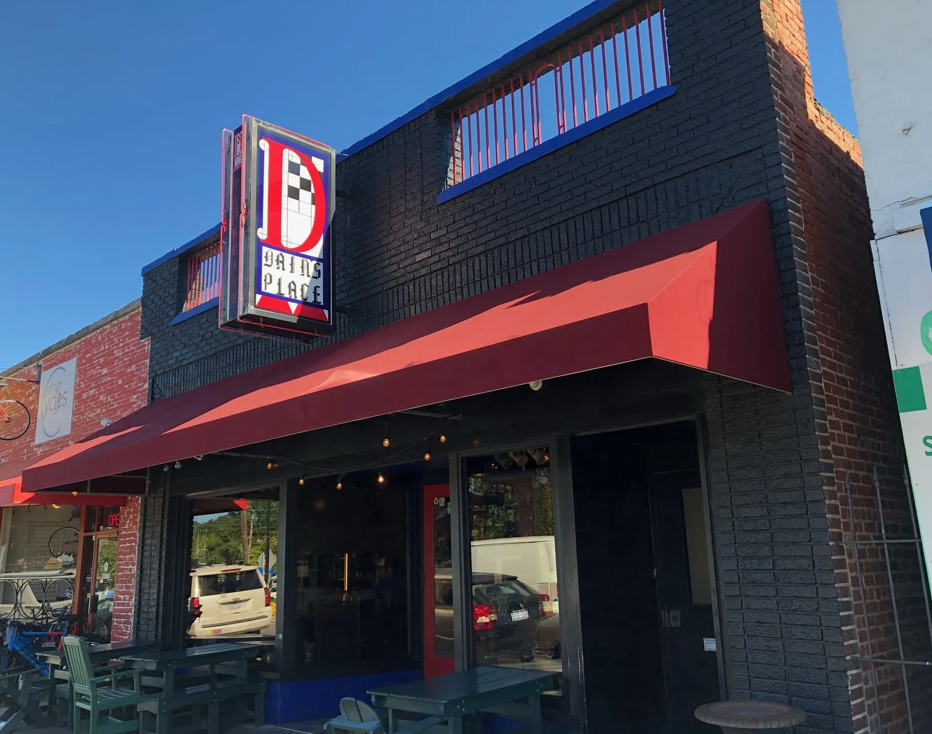 Front view of Dain's Place, a black brick building with a red awning and sunny patio.